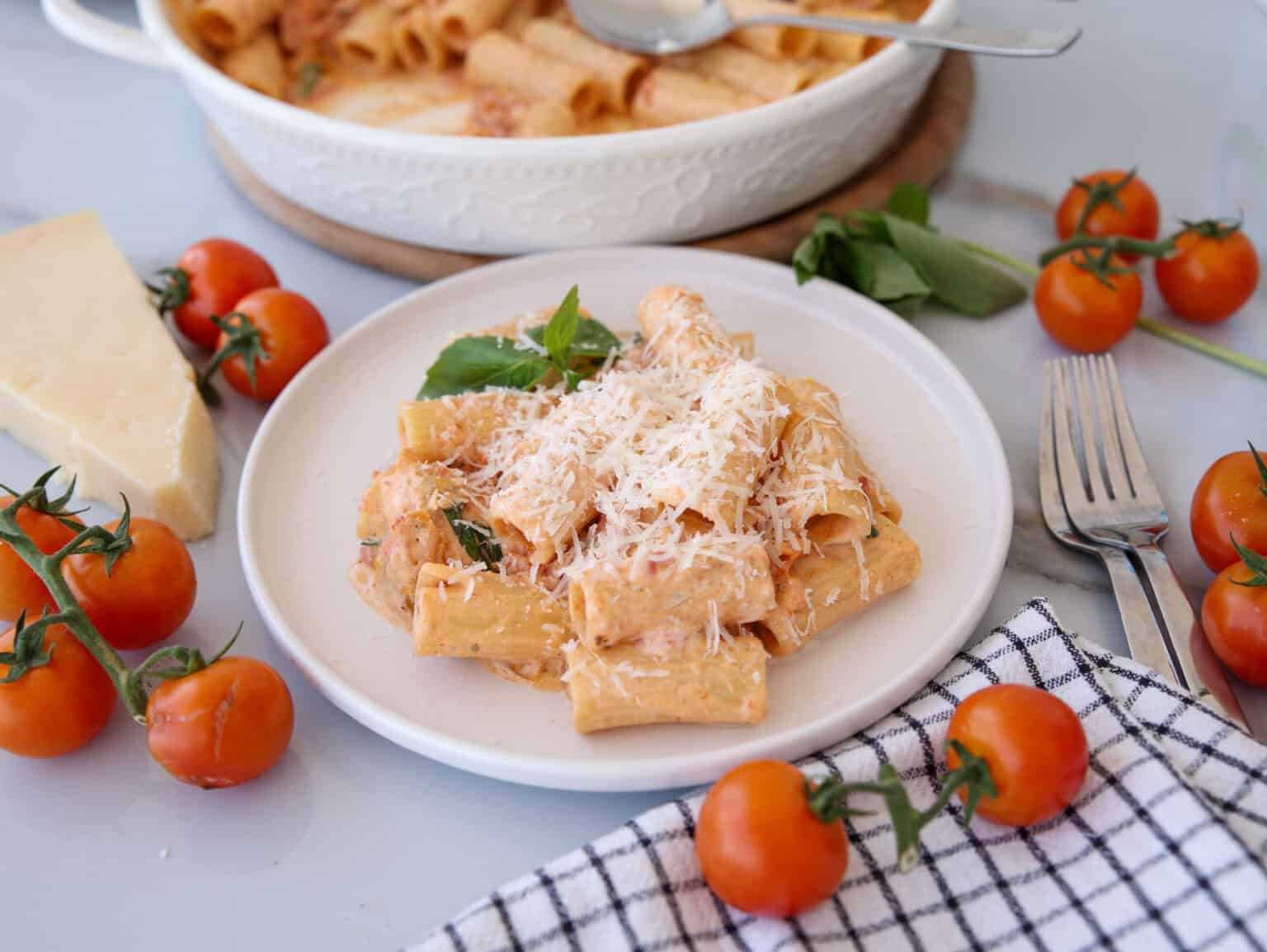 A plate of rigatoni pasta with tomato cream sauce, topped with grated cheese and basil, surrounded by fresh tomatoes, basil, a cheese wedge, cutlery, and a napkin.