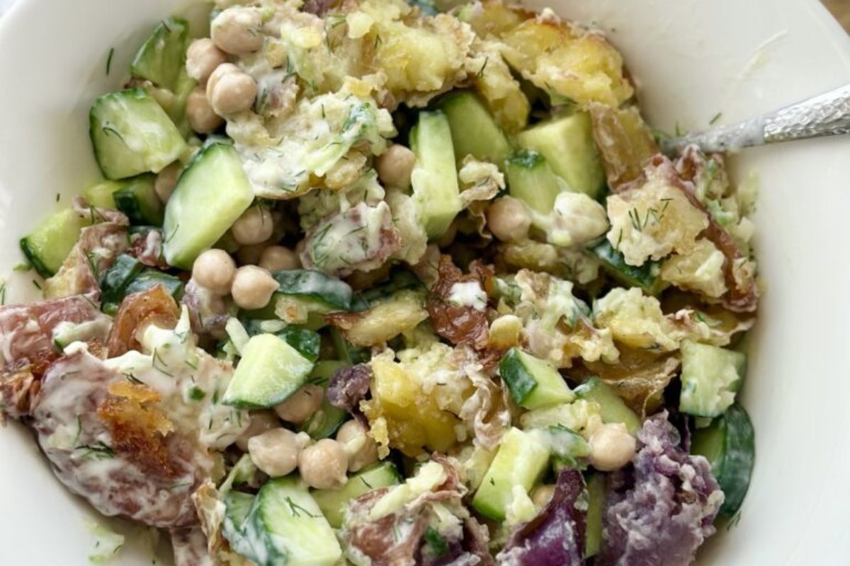 A bowl of potato salad with chickpeas, cucumber, red onion, and creamy dressing, served with a metal spoon.