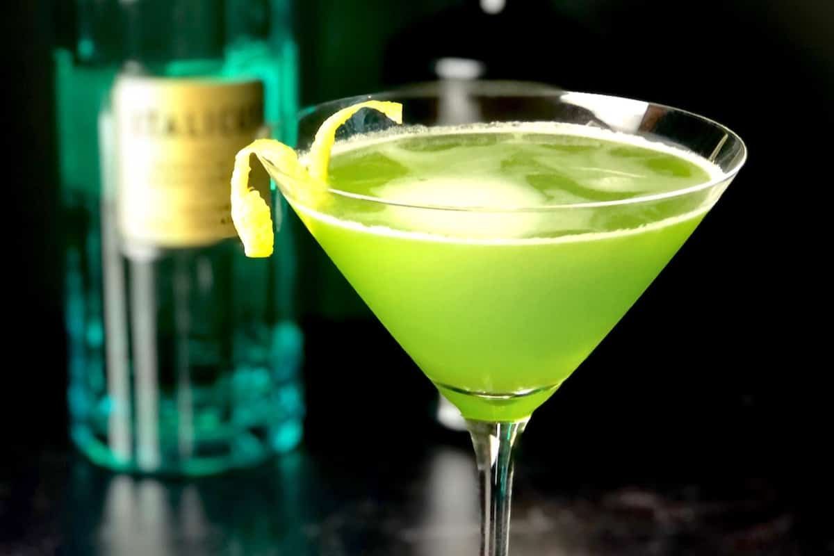 A green cocktail in a martini glass garnished with a lemon twist, with a blue bottle blurred in the background.