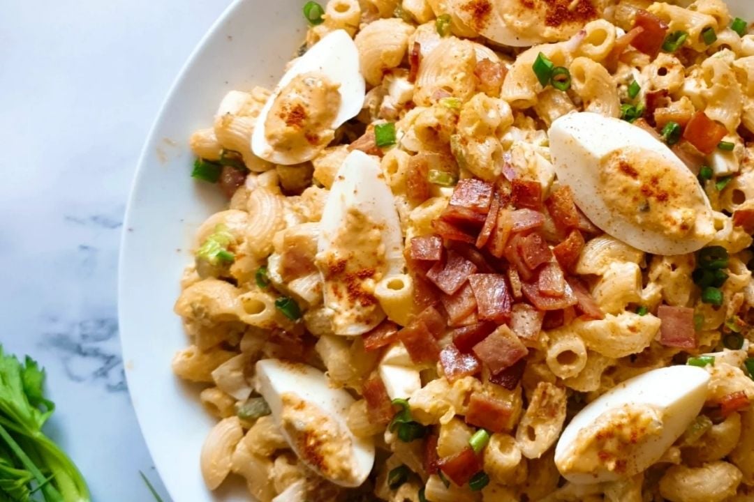 A bowl of macaroni salad topped with sliced hard-boiled eggs, diced ham, chopped green onions, and a sprinkle of paprika.