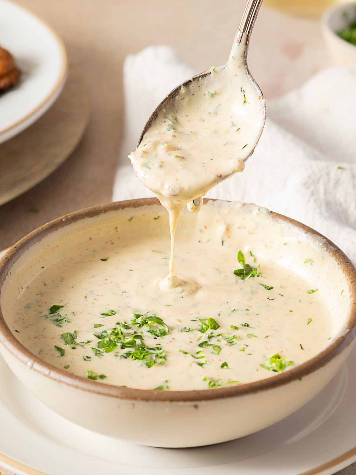 A spoon lifts creamy, herb-speckled sauce from a bowl, with fresh chopped herbs sprinkled on top.