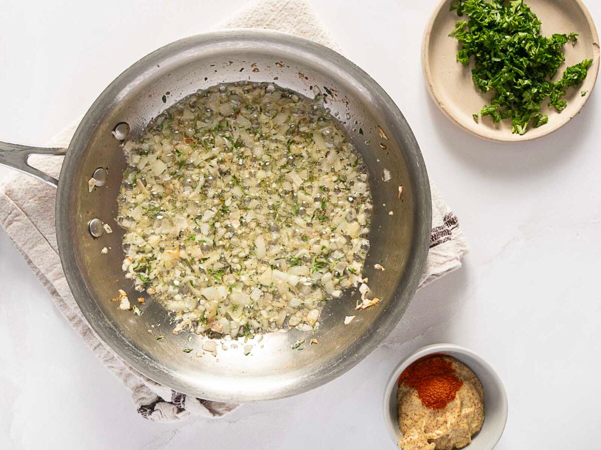 Chopped onions and herbs are being sautéed in a skillet. Nearby, a bowl of chopped parsley and a bowl with mustard and paprika are set on a white surface.