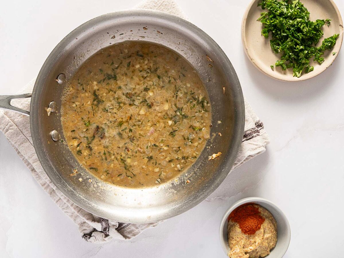 A skillet with a light brown, herb-filled sauce sits on a towel; nearby are bowls of chopped parsley and a mustard-paprika mixture.