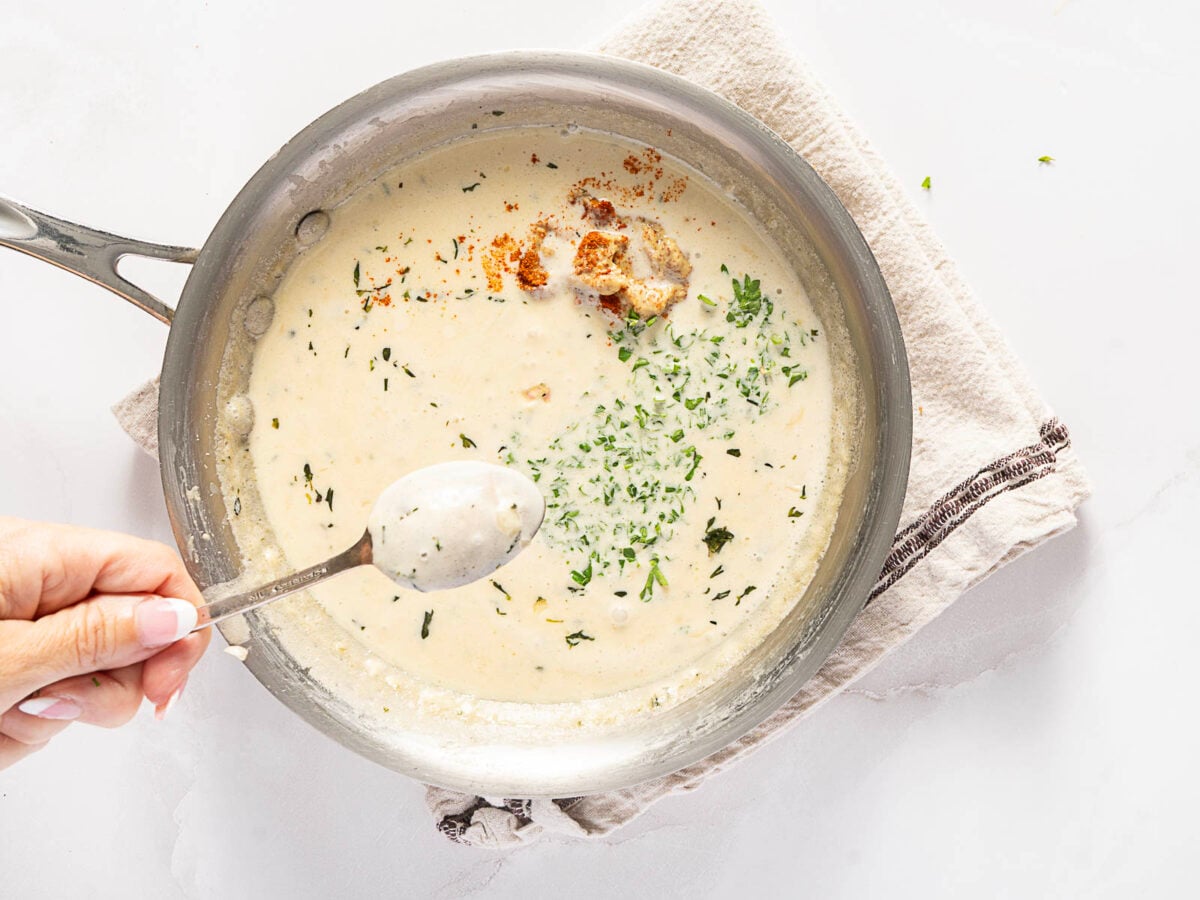 A hand holds a spoonful of creamy white soup with herbs above a pan of soup, garnished with chopped parsley and croutons, on a folded kitchen towel.