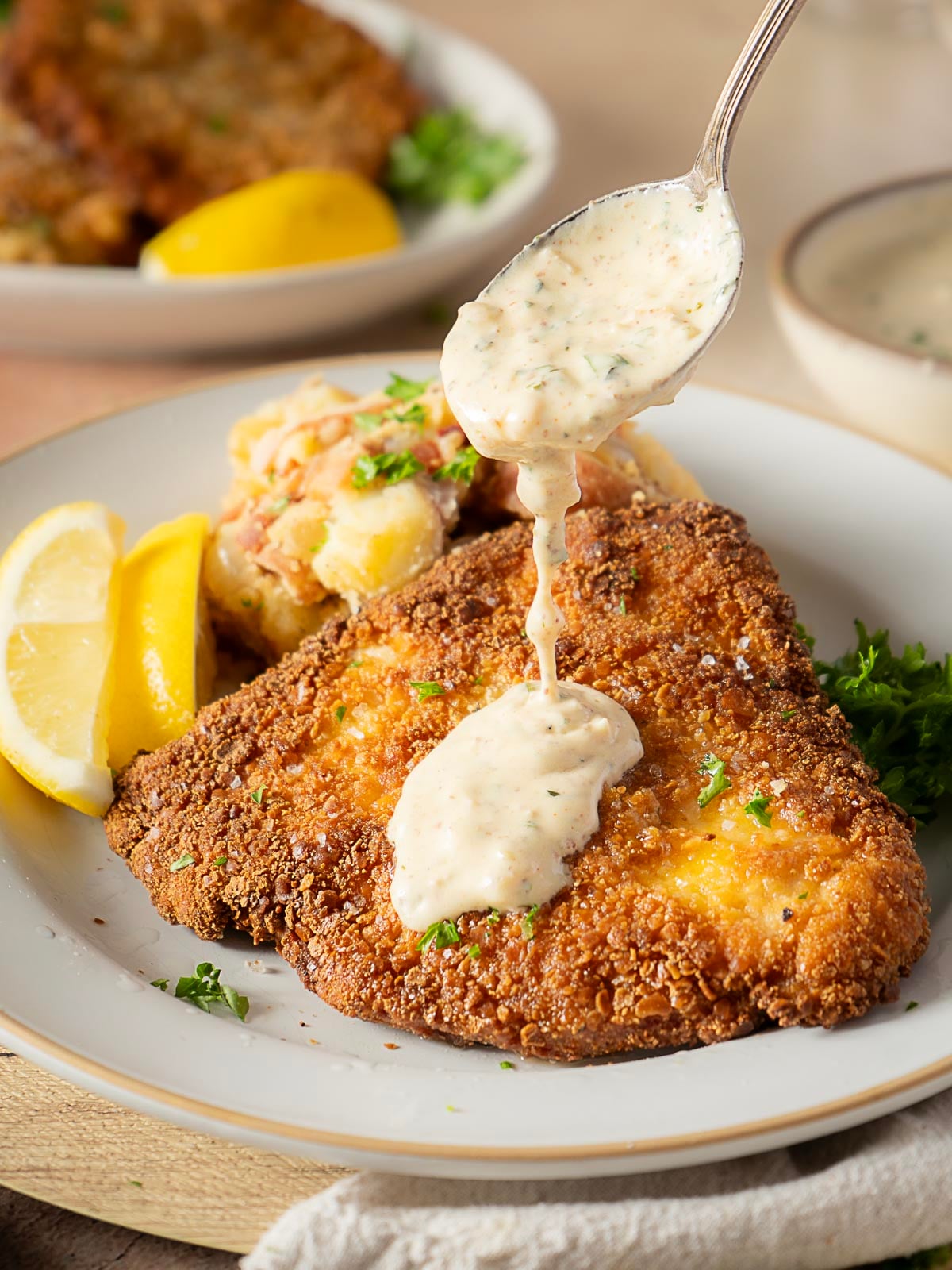 A breaded, fried fish fillet with creamy sauce being spooned on top, served with potato salad and lemon wedges on a plate.