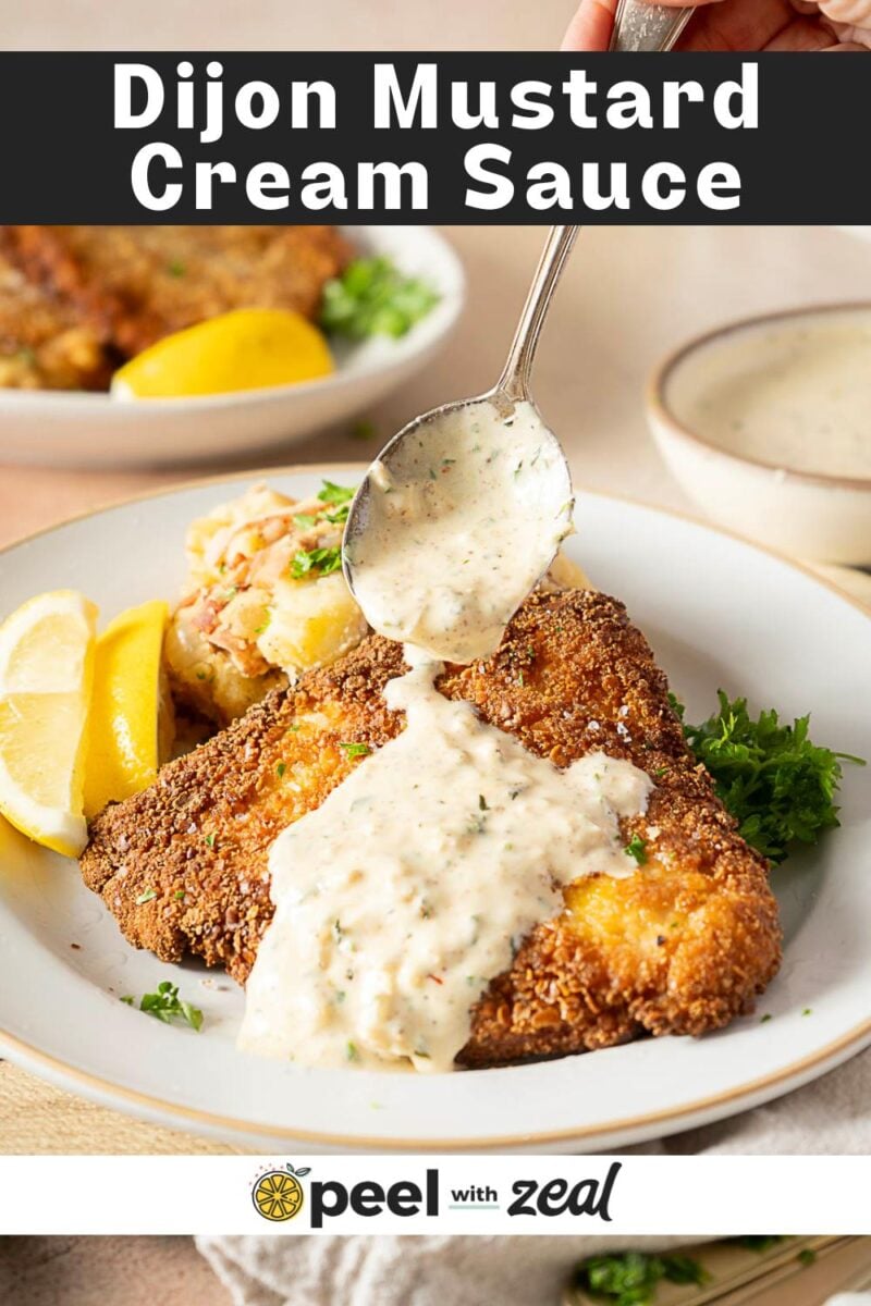 A hand spoons rich dijon cream sauce onto breaded fish on a white plate with lemon wedges, parsley, and a side dish.