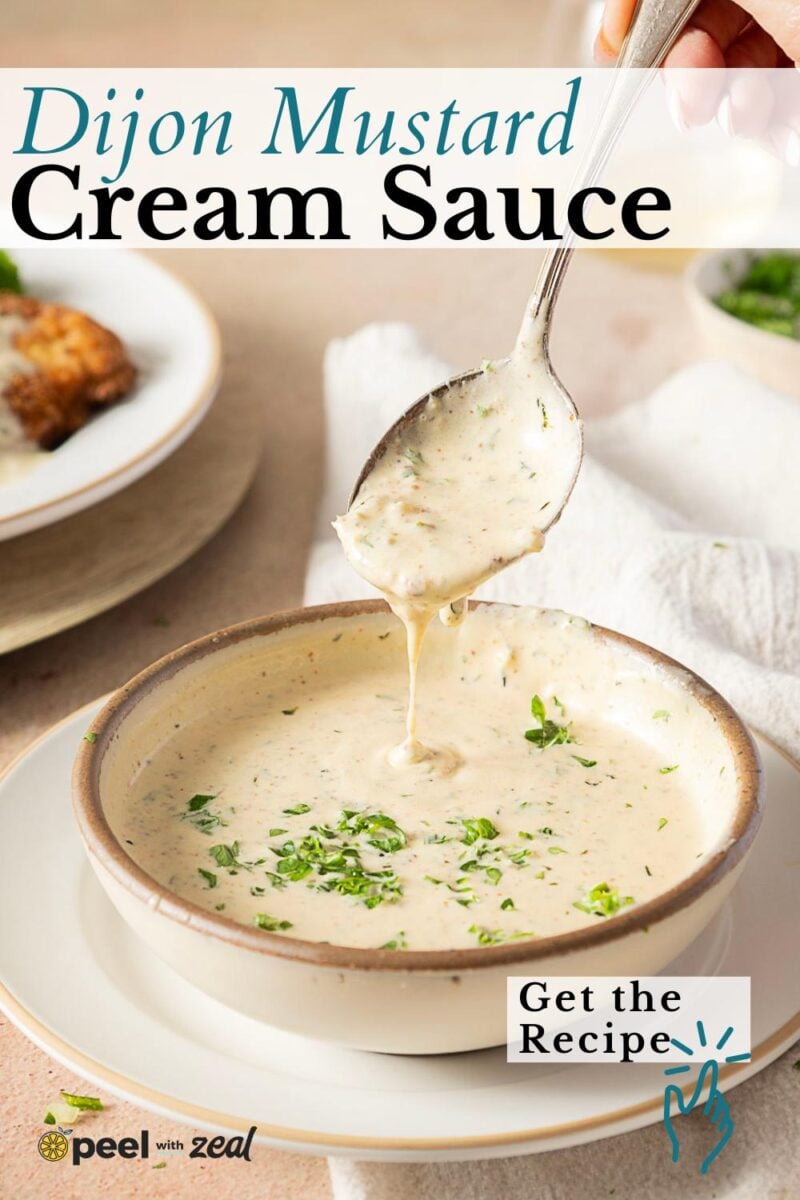 A spoon lifts luscious Dijon cream sauce from a bowl, garnished with chopped herbs. A plate of food and a white napkin are in the background.