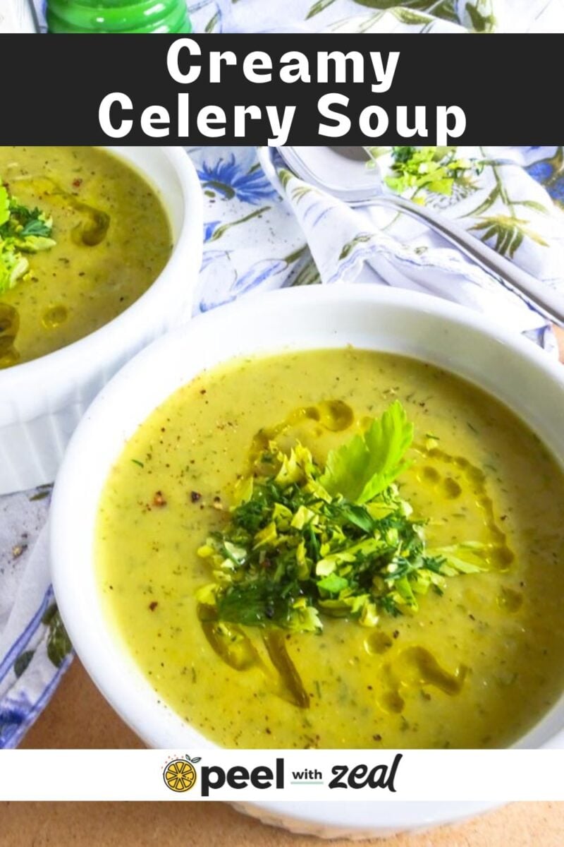Two white bowls filled with creamy celery soup, topped with chopped herbs and a drizzle of oil, placed on a patterned tablecloth with spoons and napkins in the background.