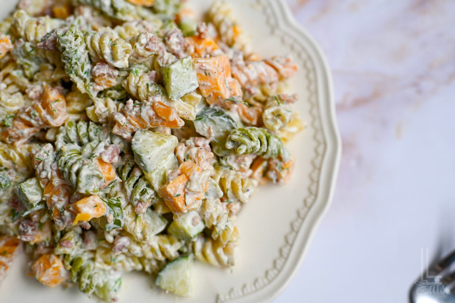 A close-up of creamy pasta salad with spiral noodles, diced cucumber, orange bell pepper, and herbs on a white plate—packed with big flavor in every bite. Perfect for those who love unique pickle recipes!.
