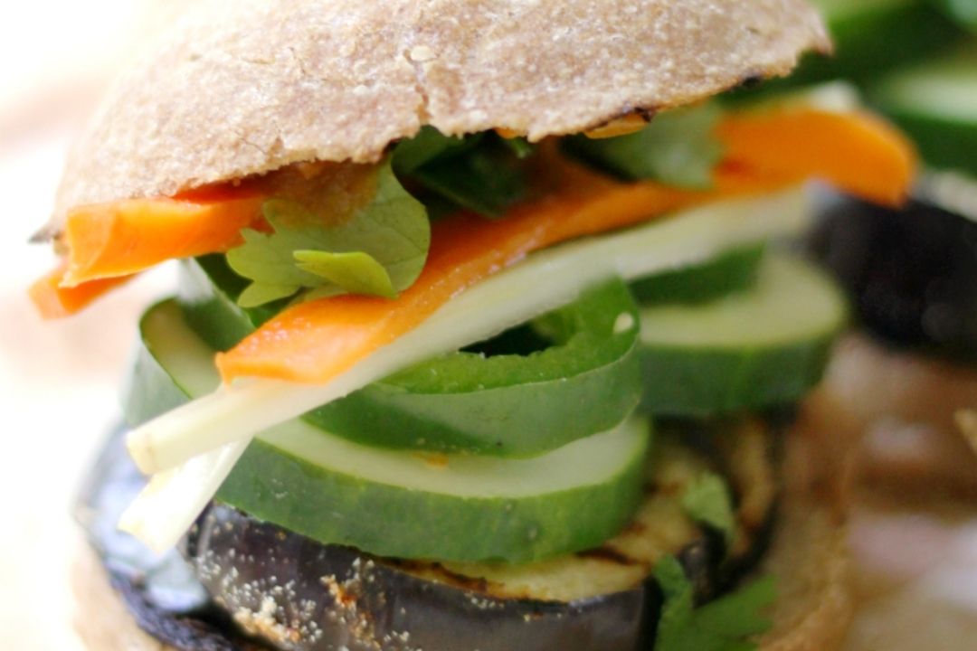 A close-up of a sandwich filled with sliced cucumber, eggplant, carrot, greens, and cheese, on whole-grain bread.