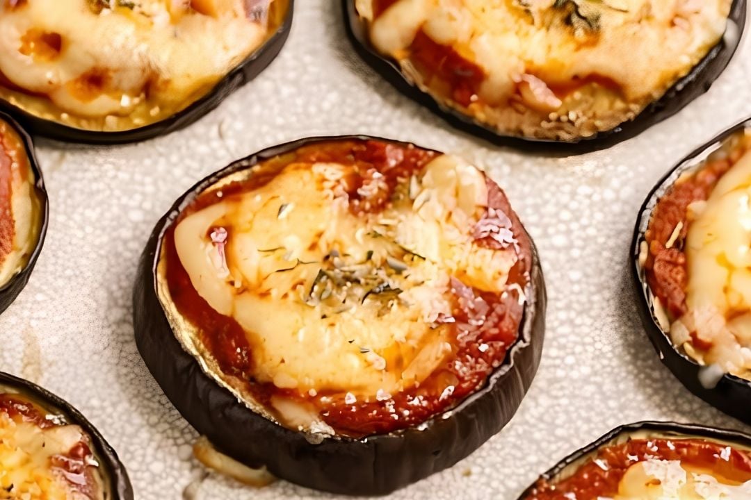 Close-up of baked eggplant slices topped with tomato sauce, melted cheese, and herbs on a textured baking sheet.