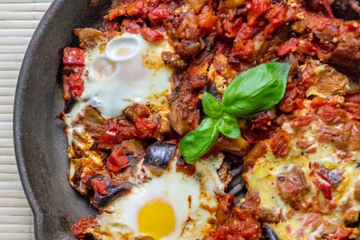 A skillet with baked eggs, tomatoes, and vegetables, garnished with fresh basil leaves.