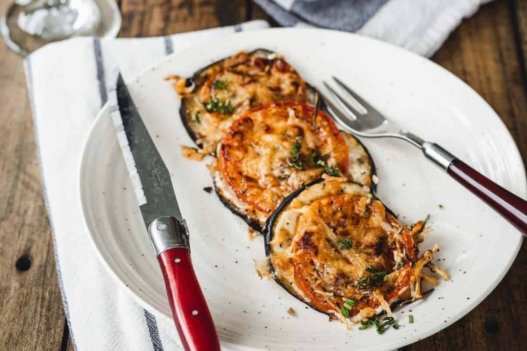 Three slices of baked eggplant topped with melted cheese and herbs are arranged on a white plate with a knife and fork beside them.