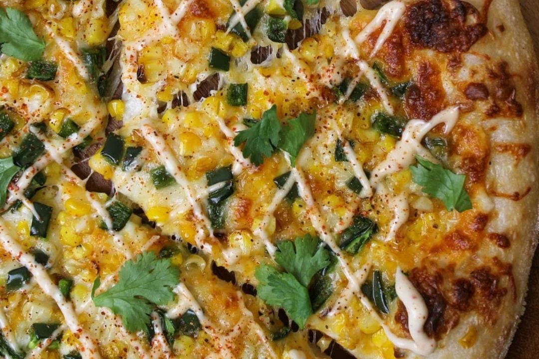 Close-up of a pizza topped with corn, chopped green peppers, melted cheese, drizzled sauce, red pepper flakes, and cilantro leaves on a golden-brown crust.
