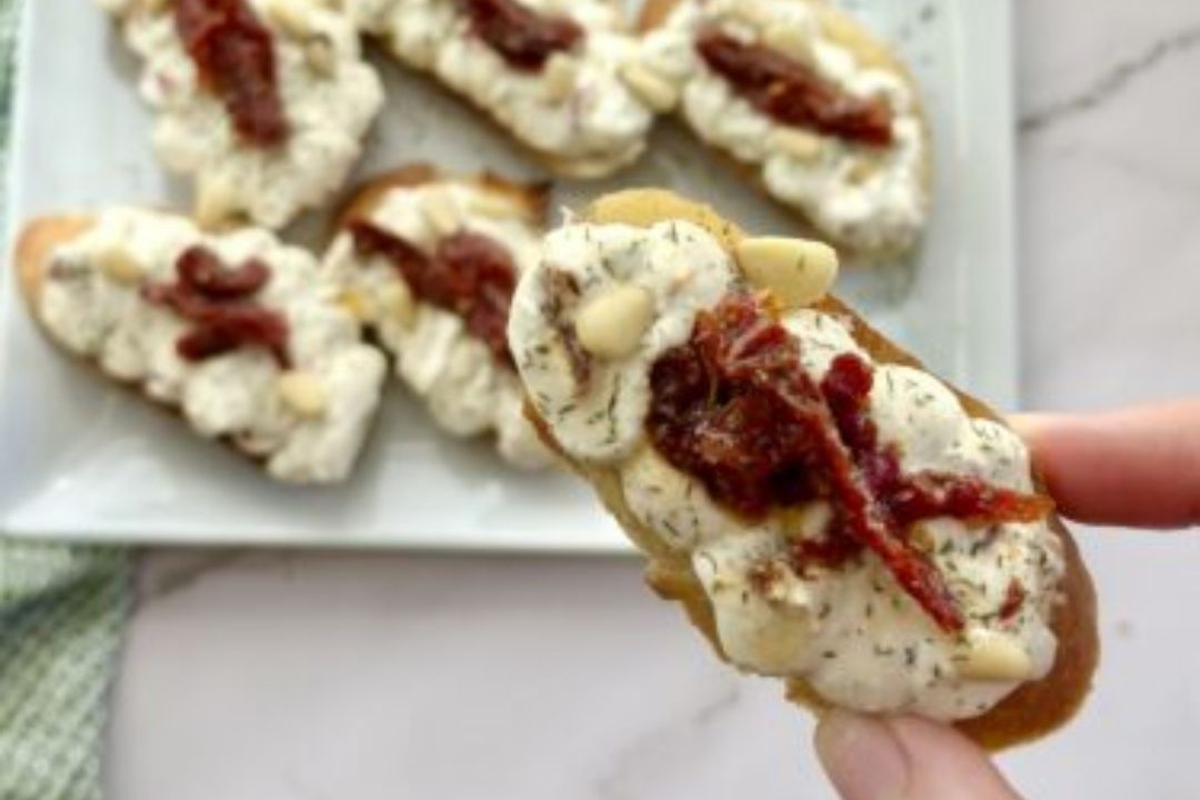 Hand holding a crostini topped with creamy spread, herbs, pine nuts, and sun-dried tomato; more crostini are arranged on a plate in the background.