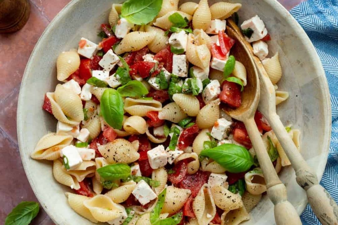 A bowl of pasta shells mixed with diced tomatoes, feta cheese, fresh basil leaves, and black pepper, with wooden serving utensils on the side.