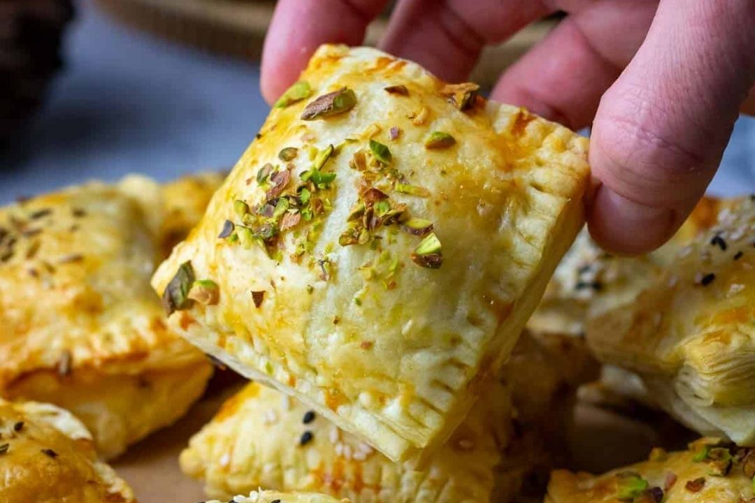 A hand holds a square puff pastry garnished with chopped nuts, with more pastries in the background.