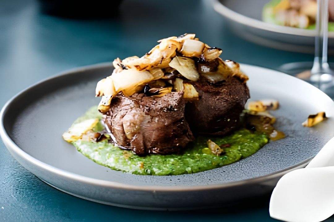 A plated steak topped with sautéed onions sits on a green vegetable puree, served on a gray plate with a blurred background.