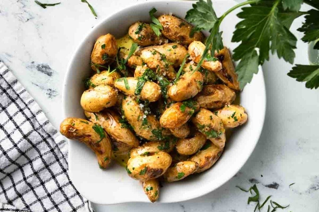 A white bowl filled with roasted fingerling potatoes garnished with chopped parsley, placed on a marble surface next to a checkered towel and a sprig of parsley.