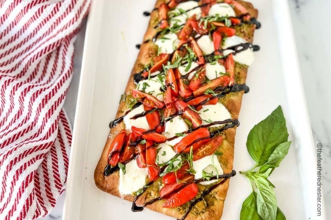 Rectangular flatbread topped with mozzarella, tomato slices, chopped basil, and balsamic glaze, served on a white tray with fresh basil leaves on the side.