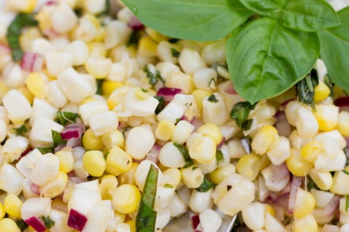 A close-up of a corn salad with chopped red onion, fresh herbs, and a basil garnish on top.