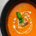 A bowl of creamy tomato soup garnished with a basil leaf, croutons, shredded cheese, black pepper, and a drizzle of cream.