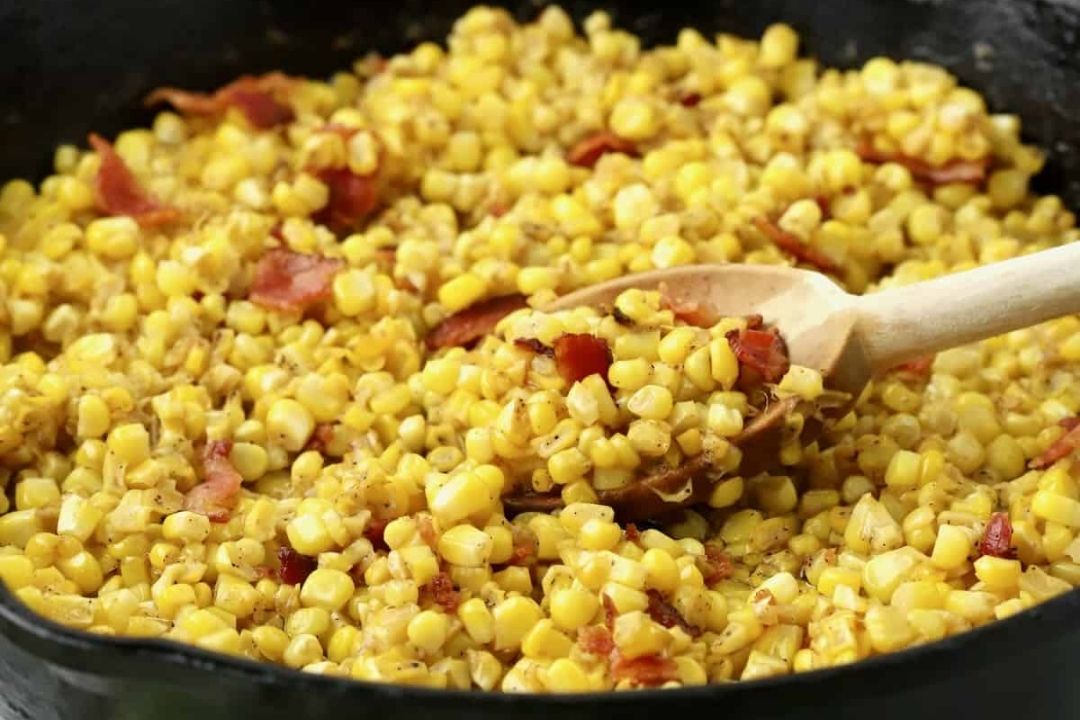 Cooked corn kernels mixed with pieces of bacon in a black skillet, with a wooden spoon stirring the mixture.