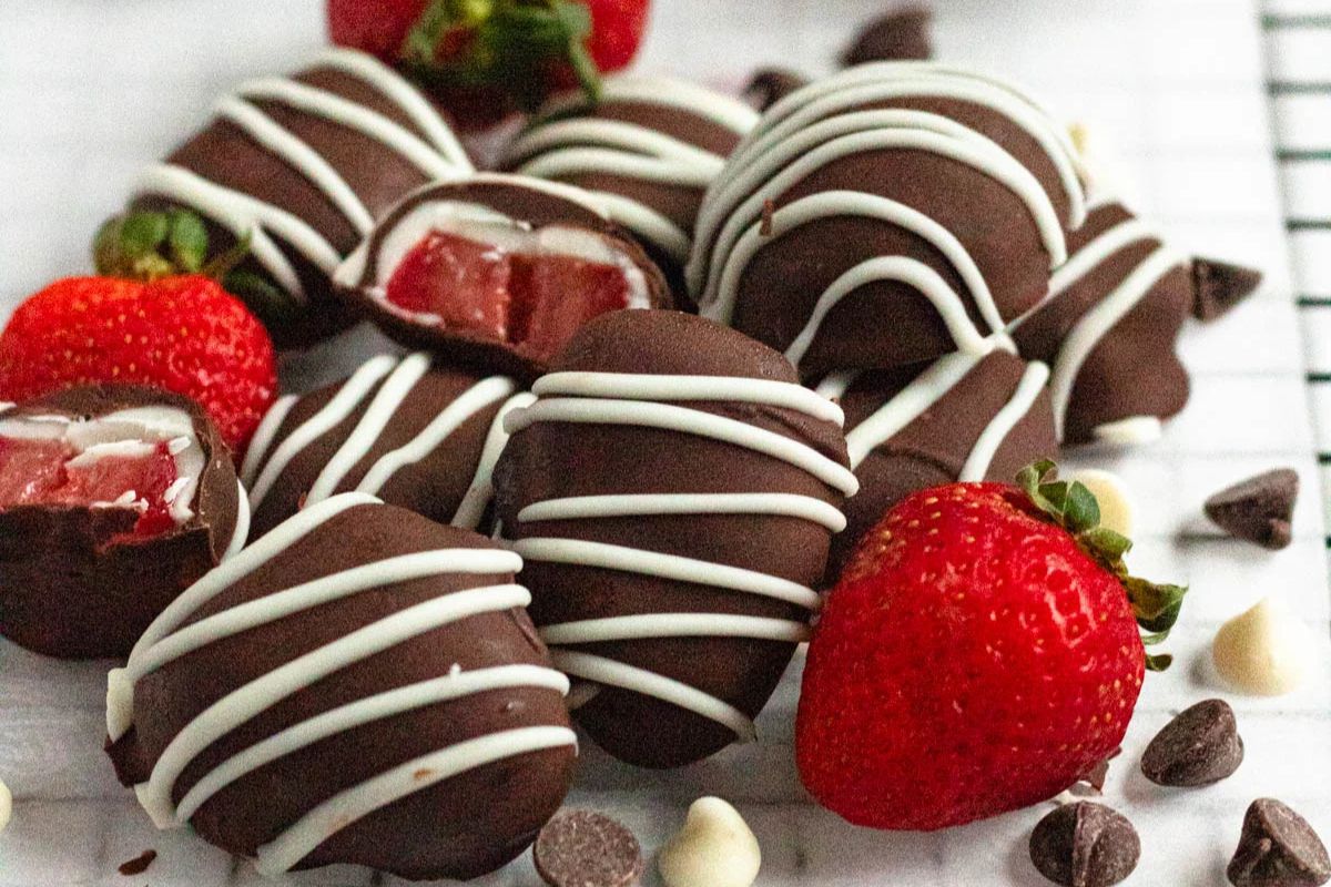 Chocolate-covered strawberry candies with white chocolate drizzle are arranged with fresh strawberries and chocolate chips on a white surface.