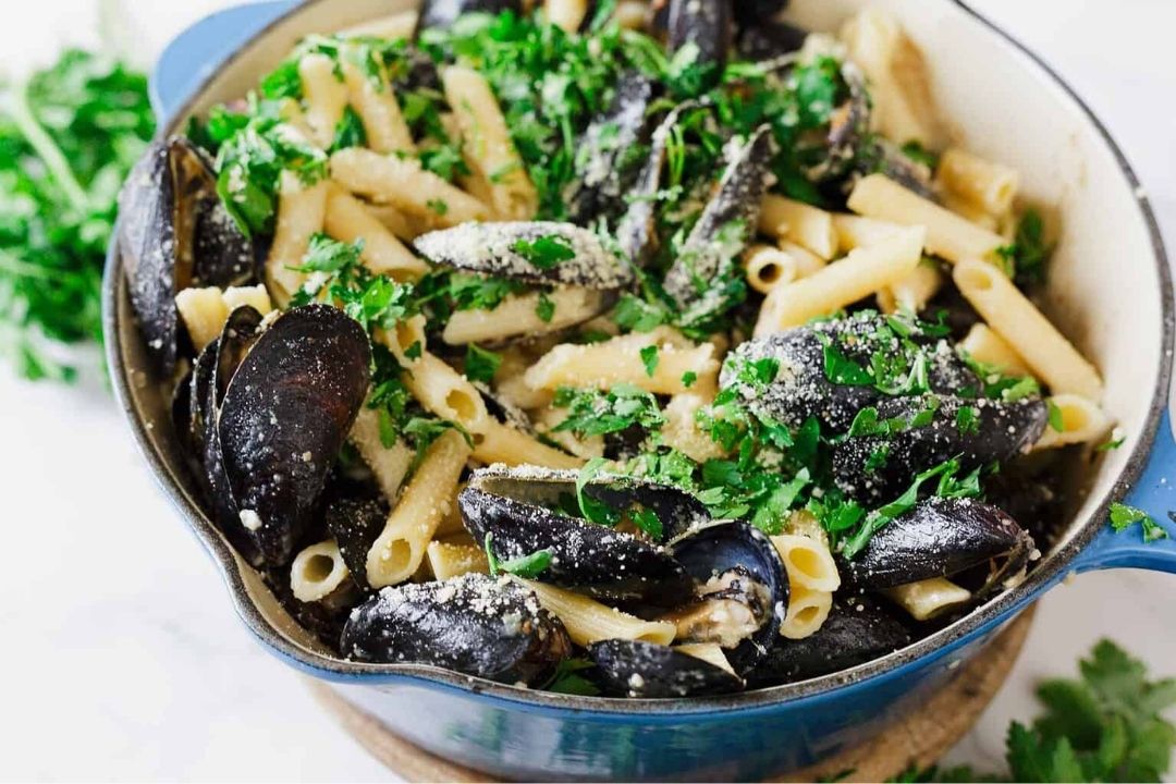 Penne pasta and mussels in a blue pot, garnished with chopped parsley and grated cheese.