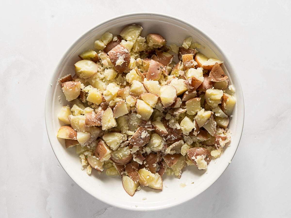 A white bowl filled with roughly chopped and partially mashed red potatoes, with skins still on, sitting on a white surface.
