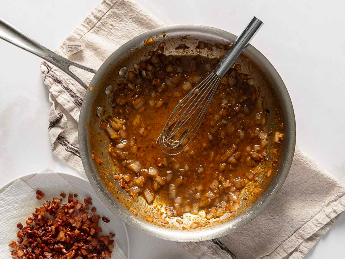 A pan with sautéed onions and brown liquid, a whisk, and a plate of cooked bacon pieces on a towel beside it.
