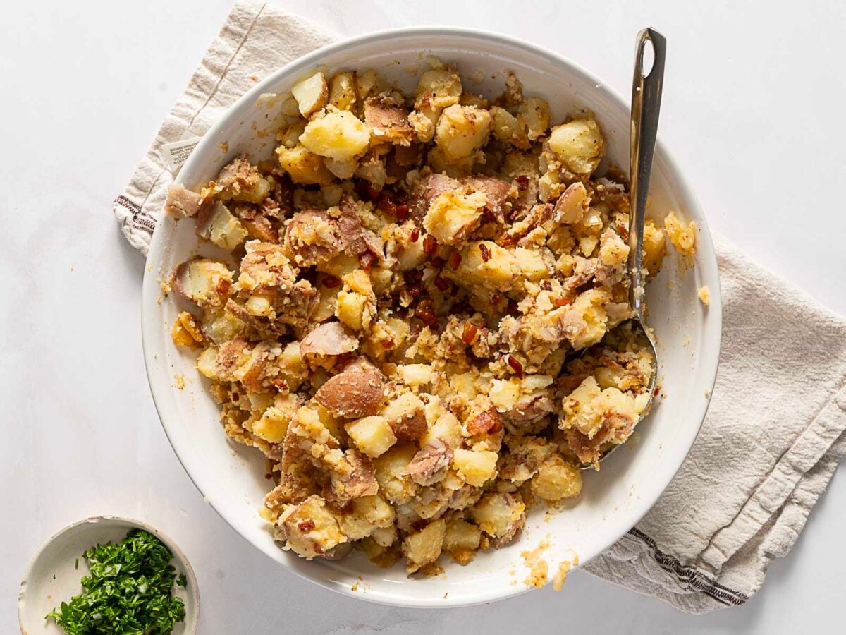 Bowl of chunky potato salad with skin-on potatoes and seasoning, placed on a white towel with a spoon and a small dish of chopped herbs nearby.
