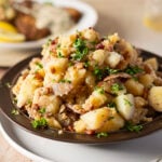 A plate of German potato salad with chopped parsley, bacon, and onions, served on a dark dish with another plate of food blurred in the background.
