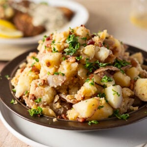 A plate of German potato salad with chopped parsley, bacon, and onions, served on a dark dish with another plate of food blurred in the background.