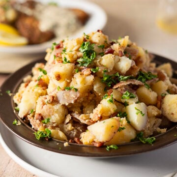 A plate of German potato salad with chopped parsley, bacon, and onions, served on a dark dish with another plate of food blurred in the background.