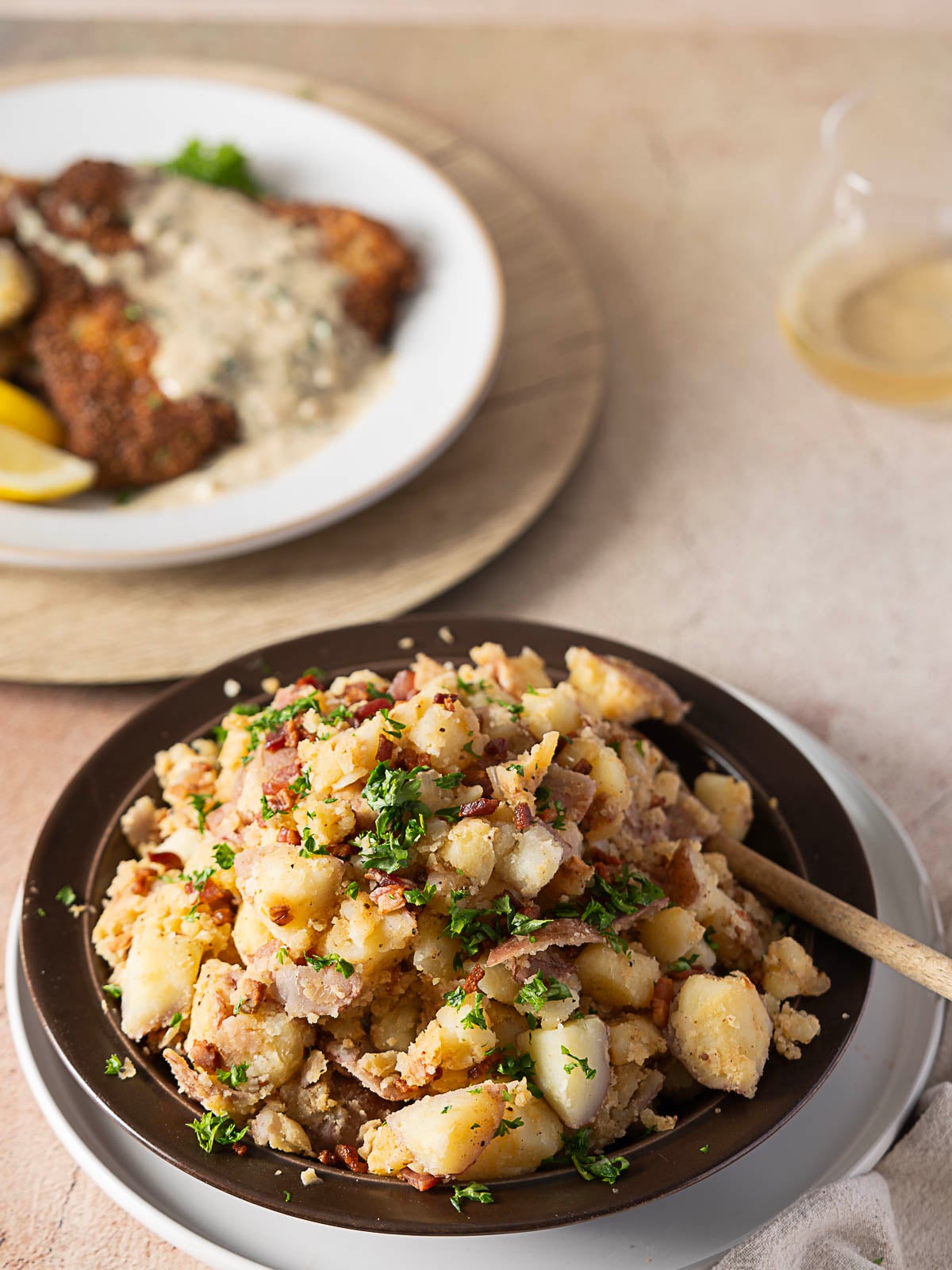 A brown plate of German potato salad with bacon and herbs sits in front of a plate of breaded meat with gravy.