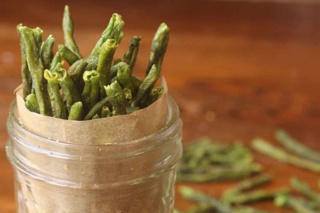 A glass jar lined with brown paper holds several crispy green bean snacks, with more green beans scattered on the wooden surface in the background.
