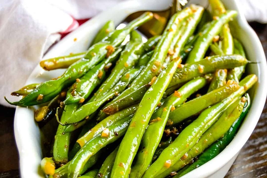 A white dish filled with cooked green beans garnished with minced garlic and sauce, placed on a wooden surface.