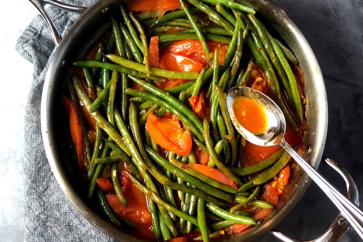 Green beans and tomato slices cooked in a red sauce in a large pan, with a spoon resting on the side.