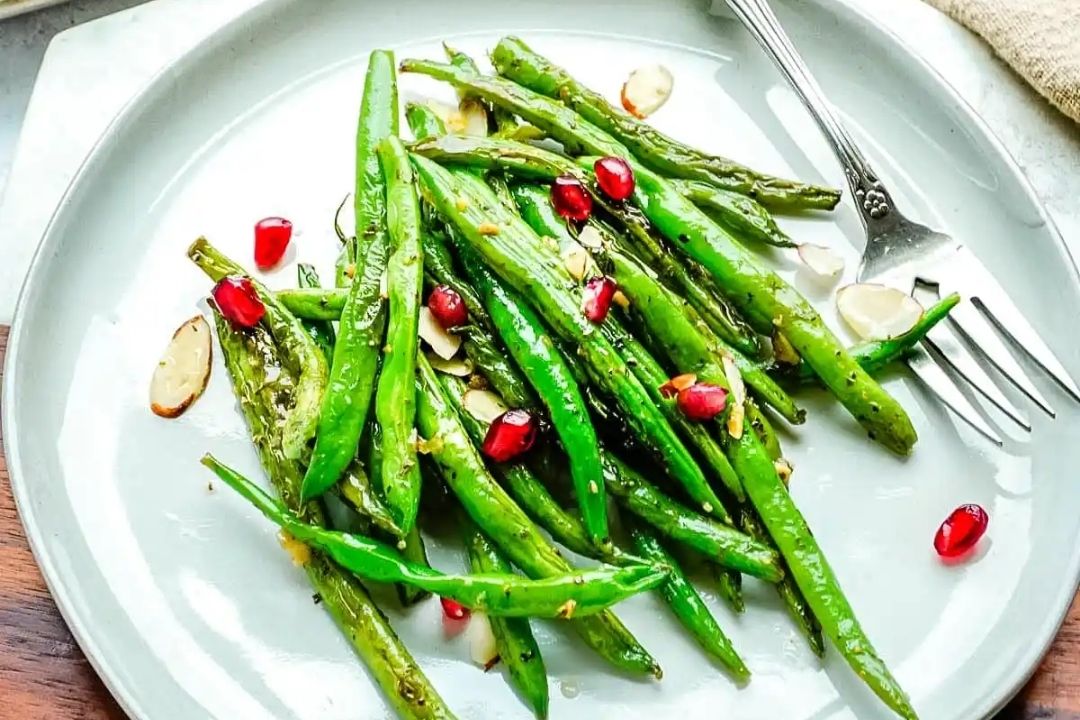 A plate of sautéed green beans garnished with sliced almonds and pomegranate seeds, with a fork on the side.