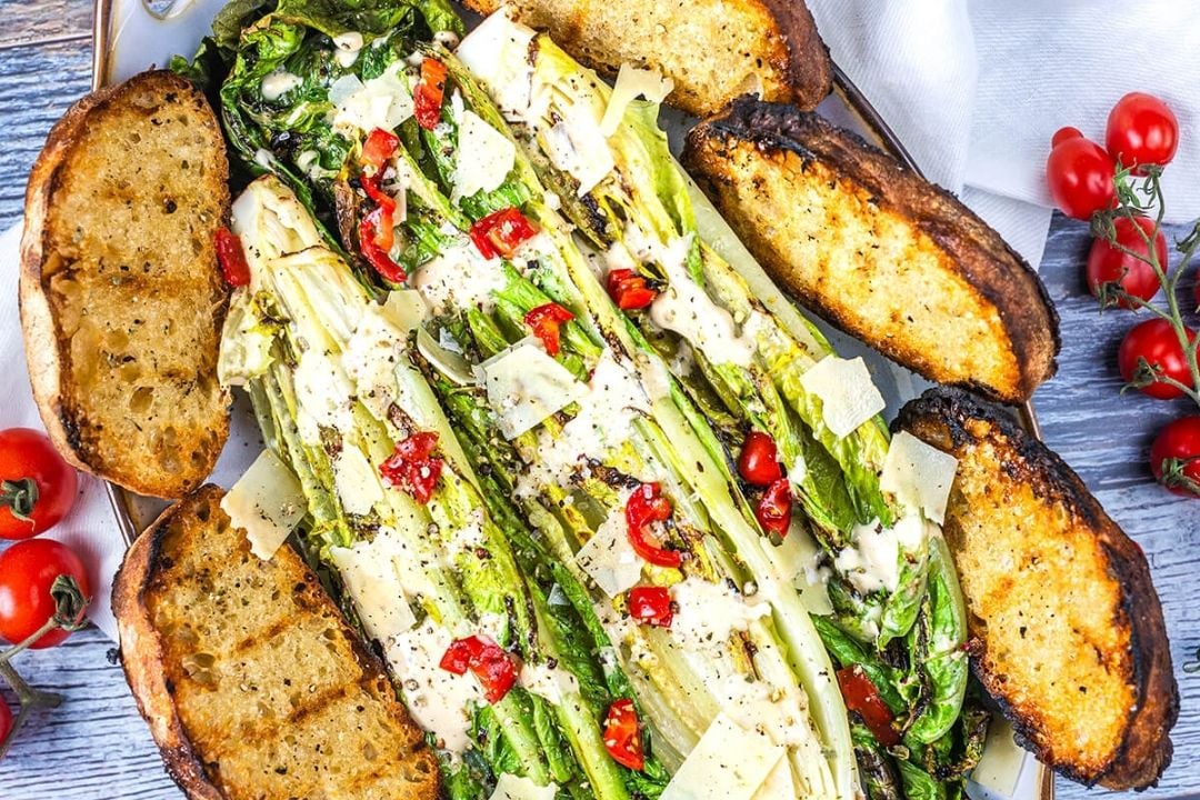 Grilled romaine lettuce topped with dressing, cherry tomatoes, and cheese, served with toasted bread slices on a plate.
