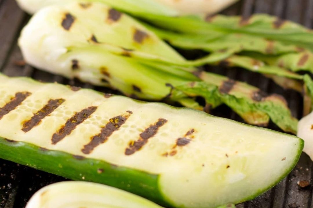 Grilled zucchini and bok choy on a barbecue grill, showing visible char marks and slight browning.
