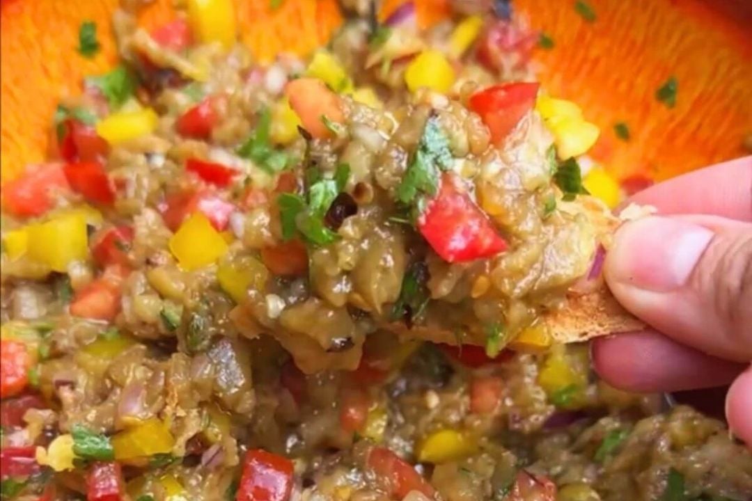 A hand holds a chip scooping up chunky dip with diced tomatoes, yellow peppers, onions, and herbs from an orange bowl.