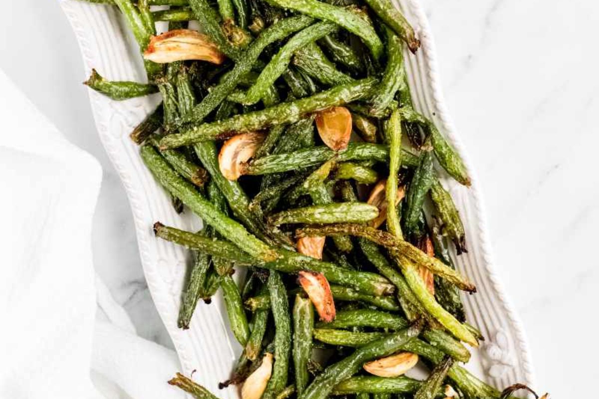 A rectangular white platter filled with roasted green beans and whole garlic cloves, viewed from above on a white surface.