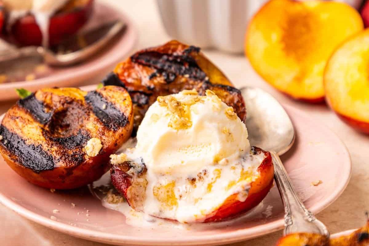 Grilled peaches topped with a scoop of vanilla ice cream and sprinkled with crumbs, served on a pink plate with a spoon.