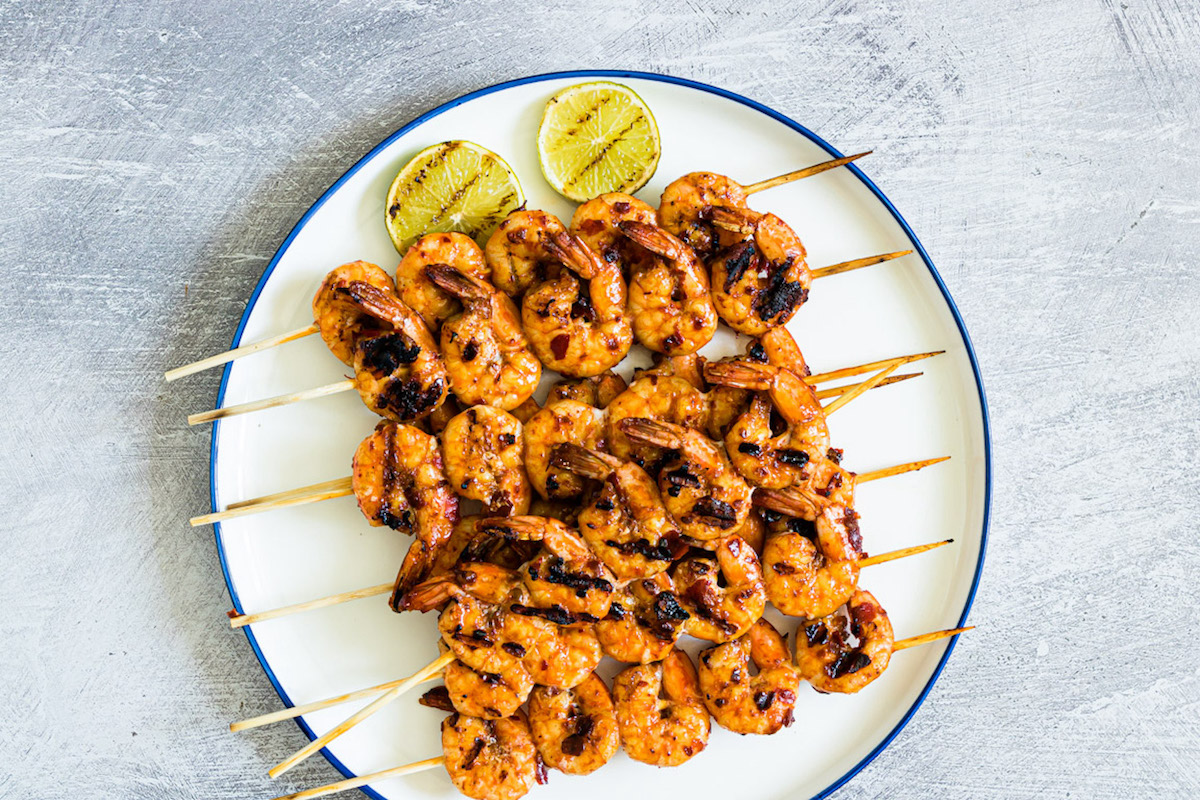 Four skewers of grilled shrimp are arranged on a white plate with two slices of grilled lime on a light textured surface.