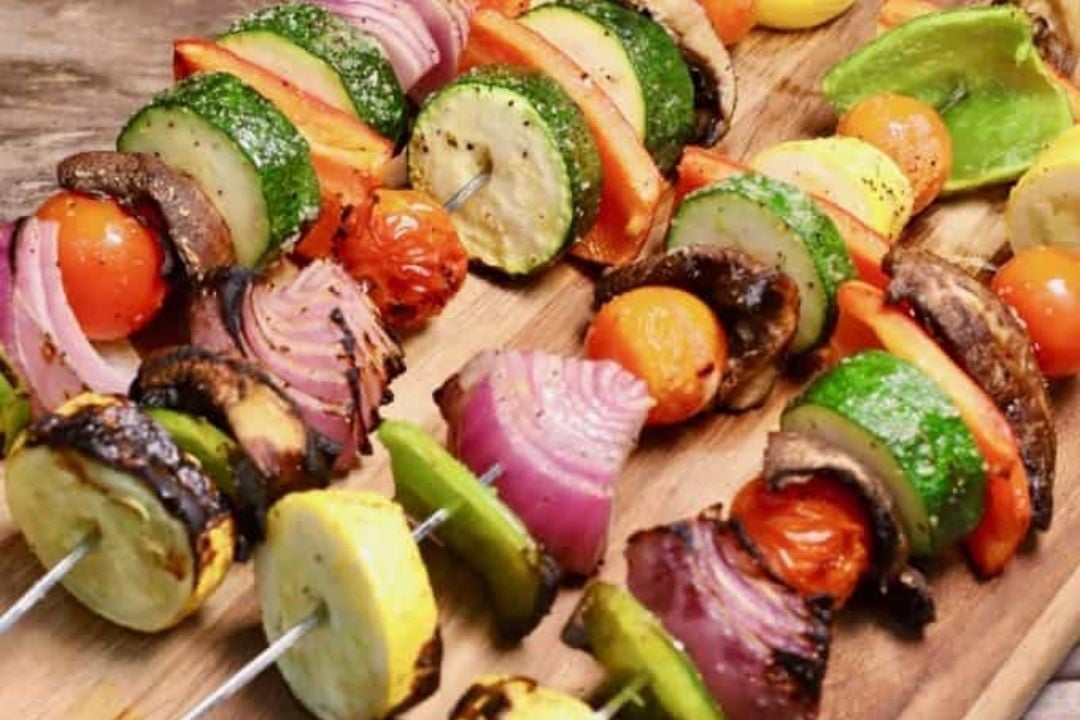 Four vegetable skewers with zucchini, cherry tomatoes, mushrooms, red onion, yellow squash, and bell peppers arranged on a wooden board.