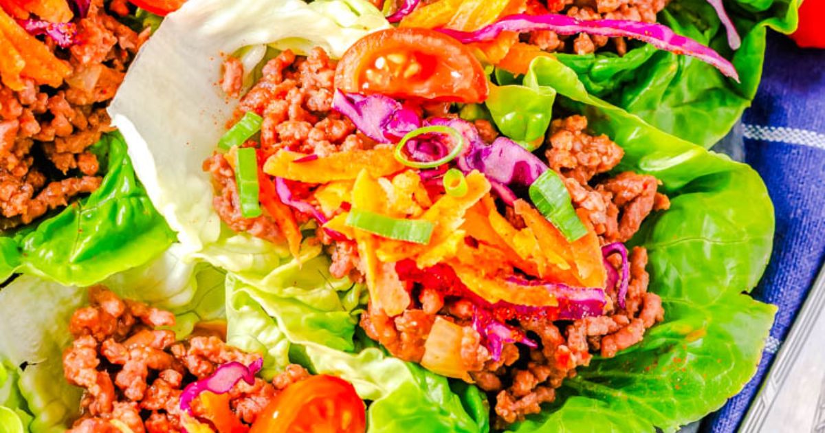 Lettuce leaves filled with seasoned ground meat, shredded carrots, sliced tomatoes, red cabbage, and green onions.
