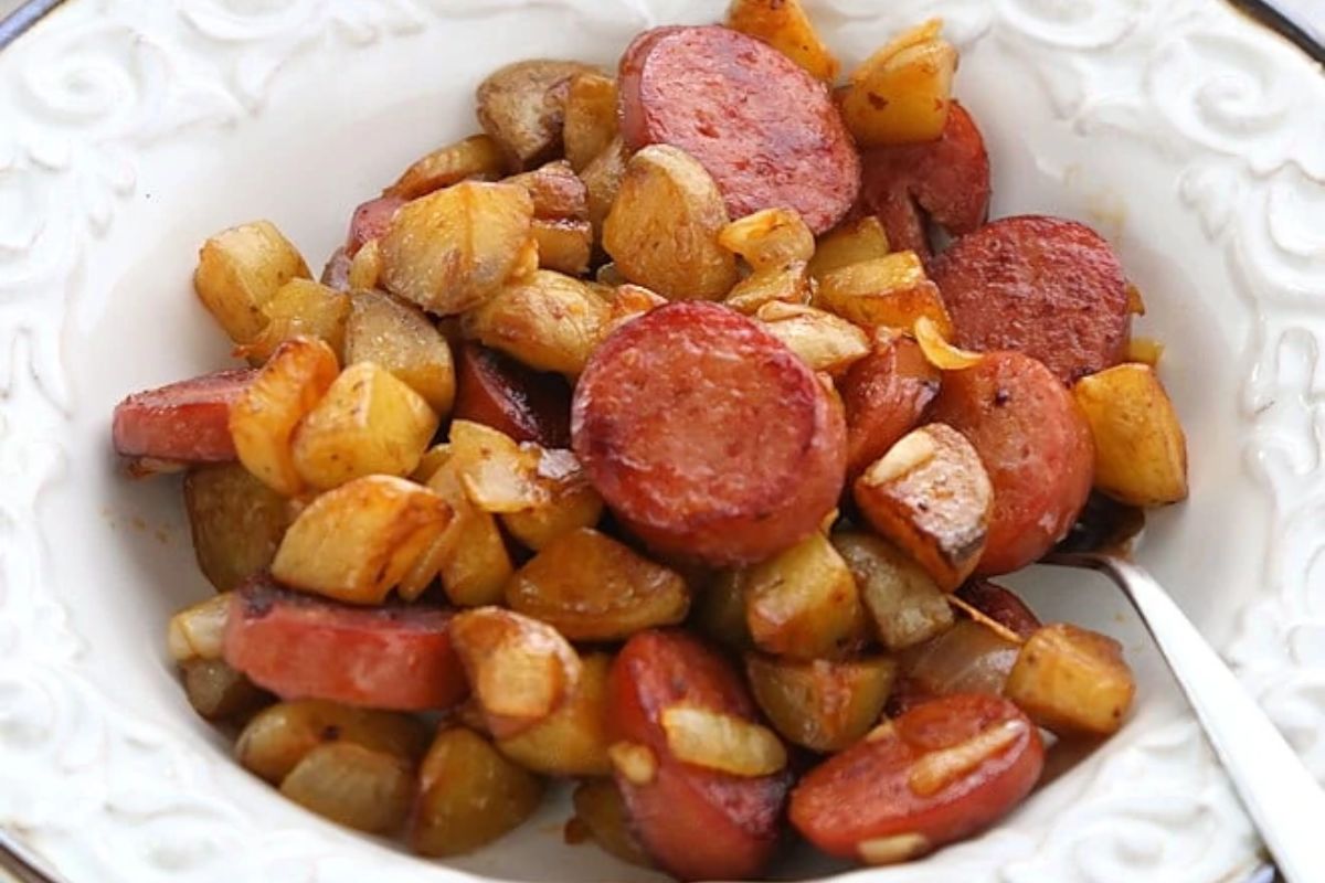 A white bowl filled with sliced sausage, diced potatoes, and onions, cooked and lightly browned, with a spoon on the side.