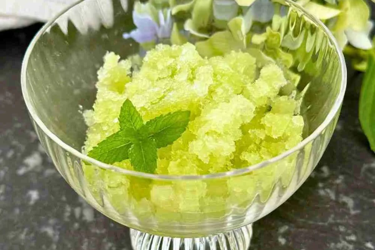 A glass dish filled with green granita topped with a fresh mint sprig, placed on a dark surface.